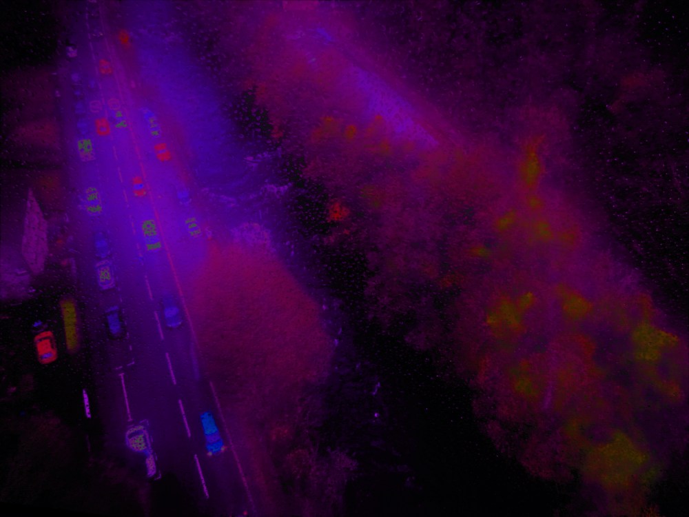 areial view of autumal foliage, a rushing river and and a busy road at dusk. The colours are blurred, jewel blues and purples with flashes of orange and red